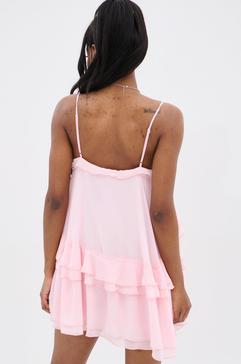 A person with long dark hair models the VANJA CHIFFON MINI DRESS in light pink, featuring a flowing design and adjustable tank straps. The elegant drape is highlighted against a plain white backdrop, emphasizing its simplicity and charm.