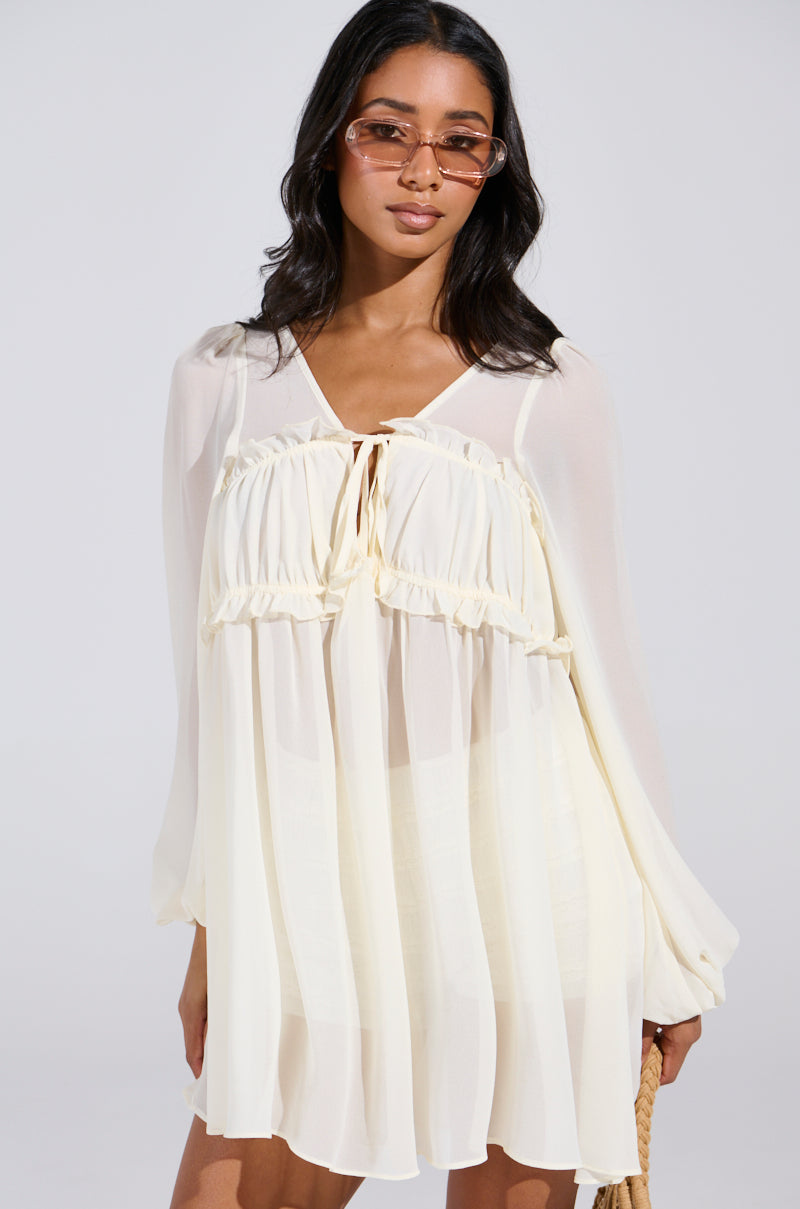 A woman exudes confidence in a light, flowy dress. Her long hair complements her sunglasses, while the ROSETTA DRESS AND BLOOMER SET boasts a loose, ruffled design with sheer fabric. The plain background perfectly highlights her stylish ensemble.