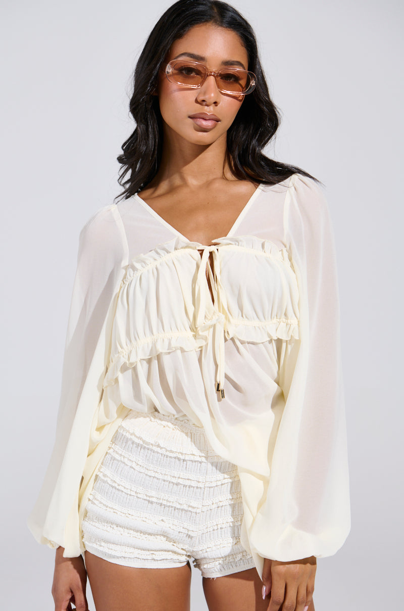 A woman is wearing the ROSETTA DRESS AND BLOOMER SET, featuring a light, flowy white blouse with ruffled details and bloomers. She has long, dark hair and sunglasses against a plain, neutral background.