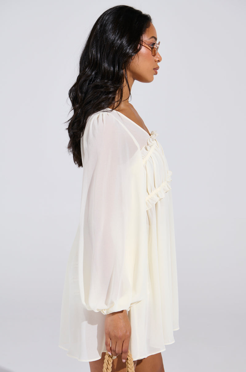 A woman with long dark hair is in profile wearing a sheer-sleeved Rosetta Dress and Bloomer Set. She holds a woven bag, gazing to the left against a plain light background that highlights her and the delicate outfit.