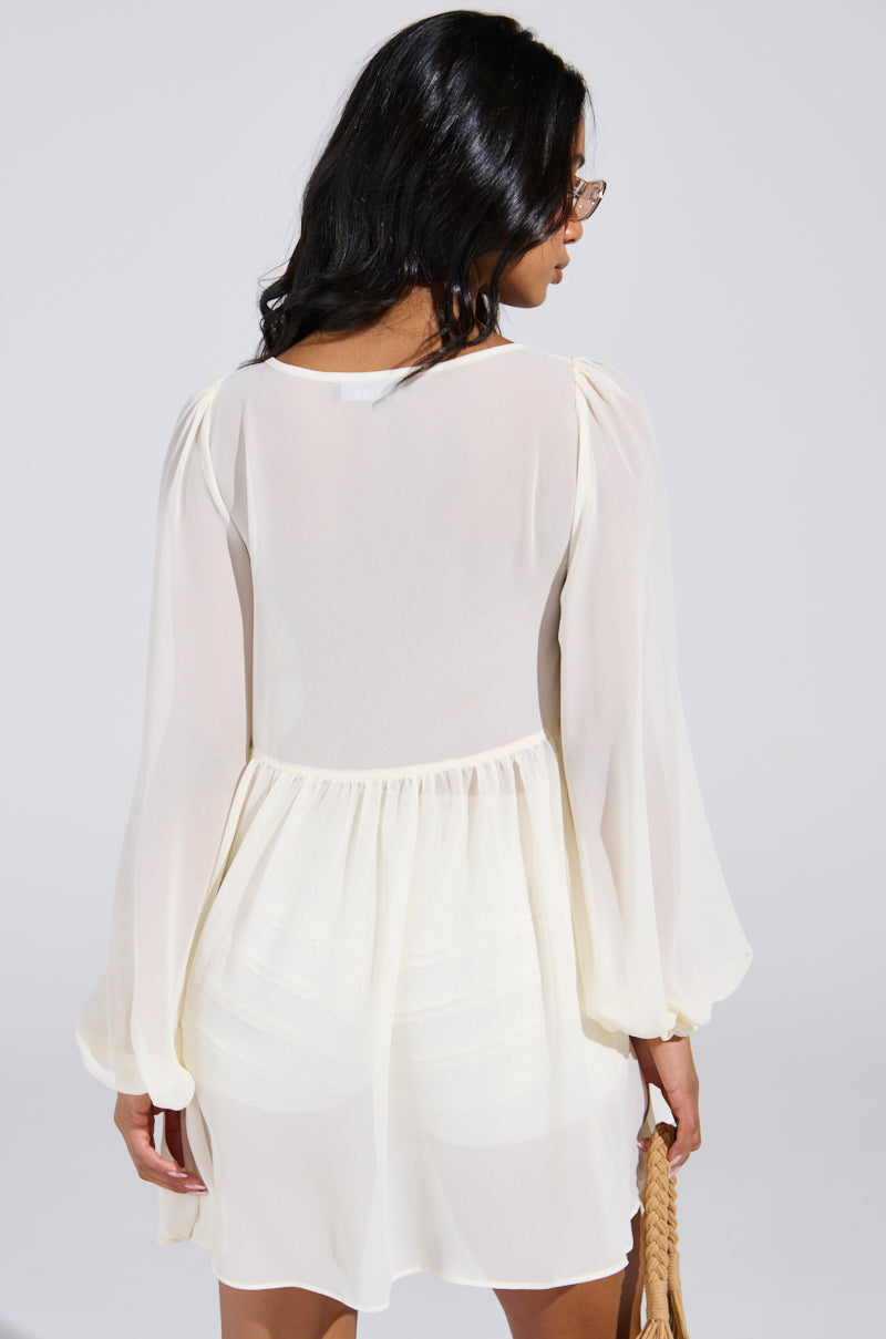 A person with long dark hair, wearing glasses, is seen from behind in a sheer Rosetta Dress and Bloomer Set with a flowy skirt, holding a woven bag. The delicate dress contrasts elegantly with the plain background.