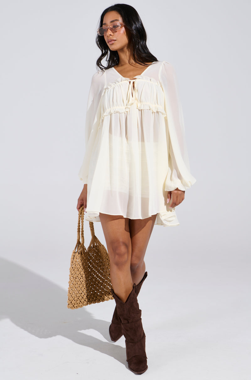 A person is walking in a flowy ROSETTA DRESS AND BLOOMER SET with sheer fabric and long sleeves, wearing sunglasses and holding a woven handbag. They're also in brown knee-high suede boots against a plain white background.