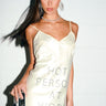 A woman with long dark hair in a high ponytail models the BUSY WORKING SATIN MINI DRESS, a cream mini dress with rhinestone "HOT PERSON AT WORK" lettering. She accessorizes with large earrings, a necklace, and holds a retro camera against a white brick wall.