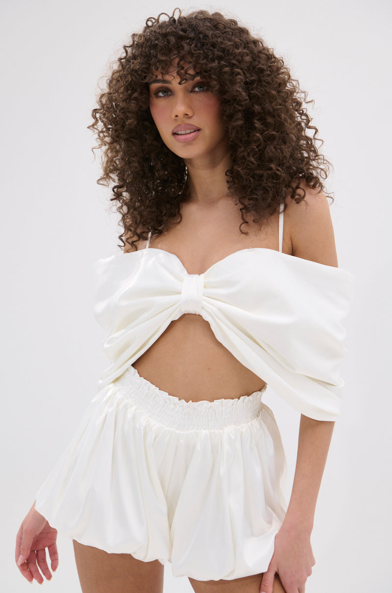 A woman with curly hair models the JULIETTE SATIN ROMPER, featuring a large bow-shaped off-the-shoulder top and matching high-waisted mini shorts, against a plain, light background.