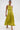 A woman confidently poses against a plain white background in the LIFE OF THE PARTY SATIN MIDI DRESS, a one-shoulder lime-green ruched satin midi with a pleated skirt and thigh-high slit, styled with gold hoop earrings and strappy gold heels.