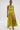 A woman stands confidently in the LIFE OF THE PARTY SATIN MIDI DRESS, a vibrant yellow-green, one-shoulder midi with ruching and a high slit. She pairs it with gold hoop earrings, an arm cuff, and gold heels against a plain white background.