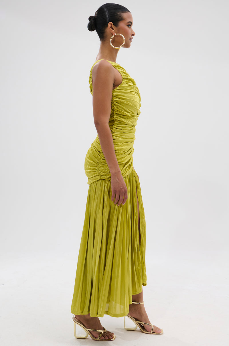 A woman stands in profile against a white background wearing the LIFE OF THE PARTY SATIN MIDI DRESS—a bright yellow-green, ruched, one-shoulder dress with a pleated skirt—styled with a bun, hoop earrings, and strappy gold heels.