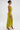A woman stands in profile against a white background wearing the LIFE OF THE PARTY SATIN MIDI DRESS—a bright yellow-green, ruched, one-shoulder dress with a pleated skirt—styled with a bun, hoop earrings, and strappy gold heels.