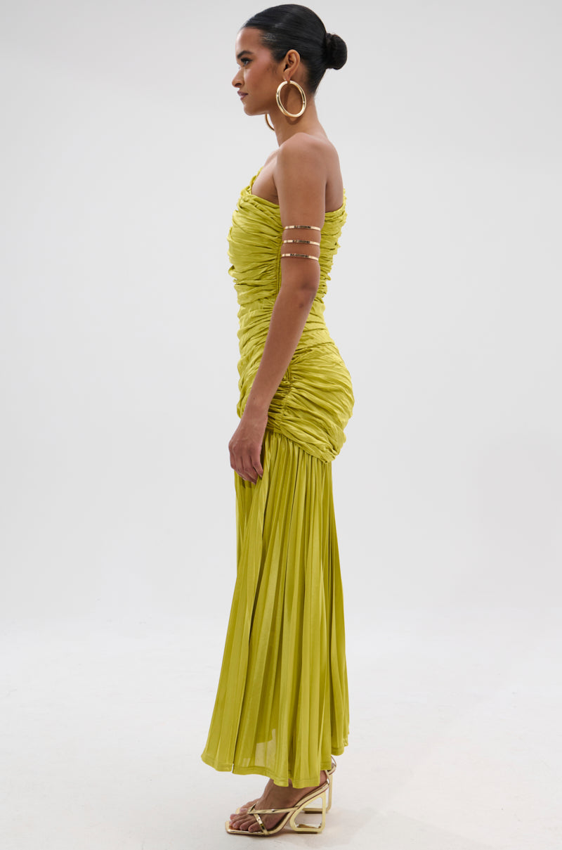 A woman stands in profile against a plain background, wearing the LIFE OF THE PARTY SATIN MIDI DRESS with ruched yellow-green fabric, pleated skirt, gold arm cuffs, large hoop earrings, heeled sandals, and a sleek bun.