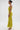 A woman stands in profile against a plain background, wearing the LIFE OF THE PARTY SATIN MIDI DRESS with ruched yellow-green fabric, pleated skirt, gold arm cuffs, large hoop earrings, heeled sandals, and a sleek bun.