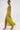A woman walks confidently in the LIFE OF THE PARTY SATIN MIDI DRESS—a bright yellow, ruched midi dress with a high slit—paired with gold heels and large hoop earrings against a plain white background.