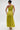 A woman stands facing away, wearing the LIFE OF THE PARTY SATIN MIDI DRESS in lime green with a ruched bodice and flowing skirt. She styles it with a bun, gold hoop earrings, an arm cuff, and high-heeled sandals against a plain background.