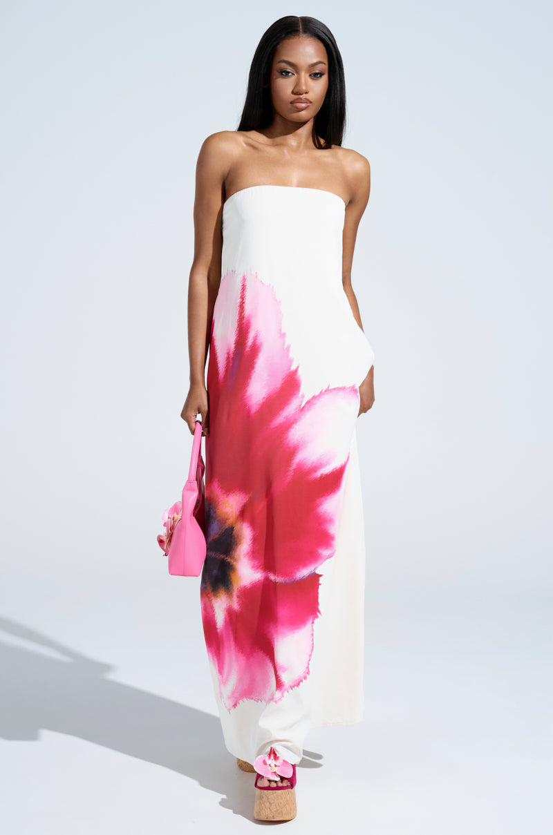A woman exudes confidence in the SWEET AND SPICY SATIN MAXI DRESS, a strapless white maxi featuring bold pink and red floral prints. She pairs it with a pink bag and platform sandals, posed against a simple, well-lit background.