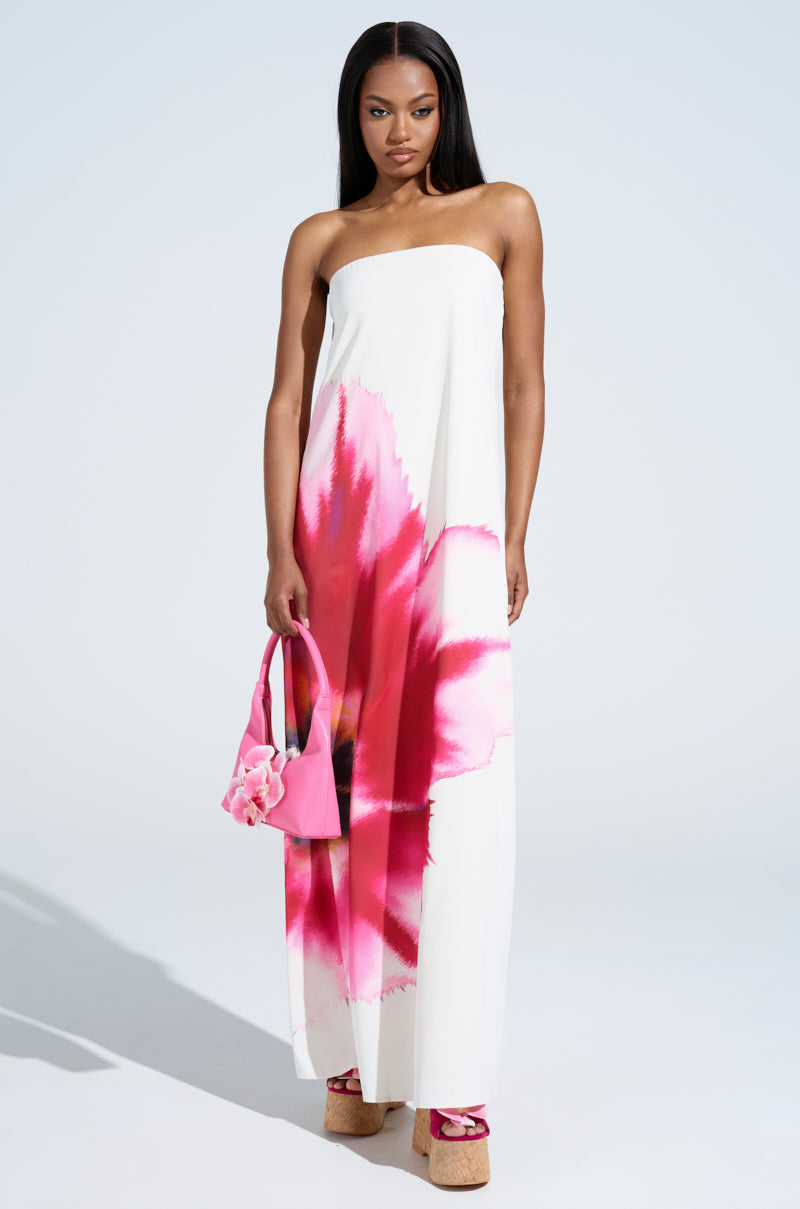A woman in the SWEET AND SPICY SATIN MAXI DRESS—a strapless maxi with bold pink florals—holds a pink handbag and wears pink platform sandals, standing against a light background.