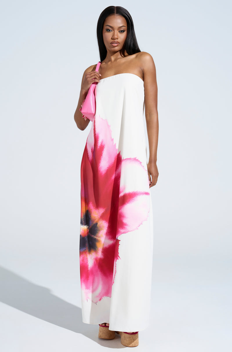 A woman wearing the SWEET AND SPICY SATIN MAXI DRESS, featuring a strapless silhouette and vibrant pink and red floral print, holds a pink purse and stands in platform sandals against a light background.