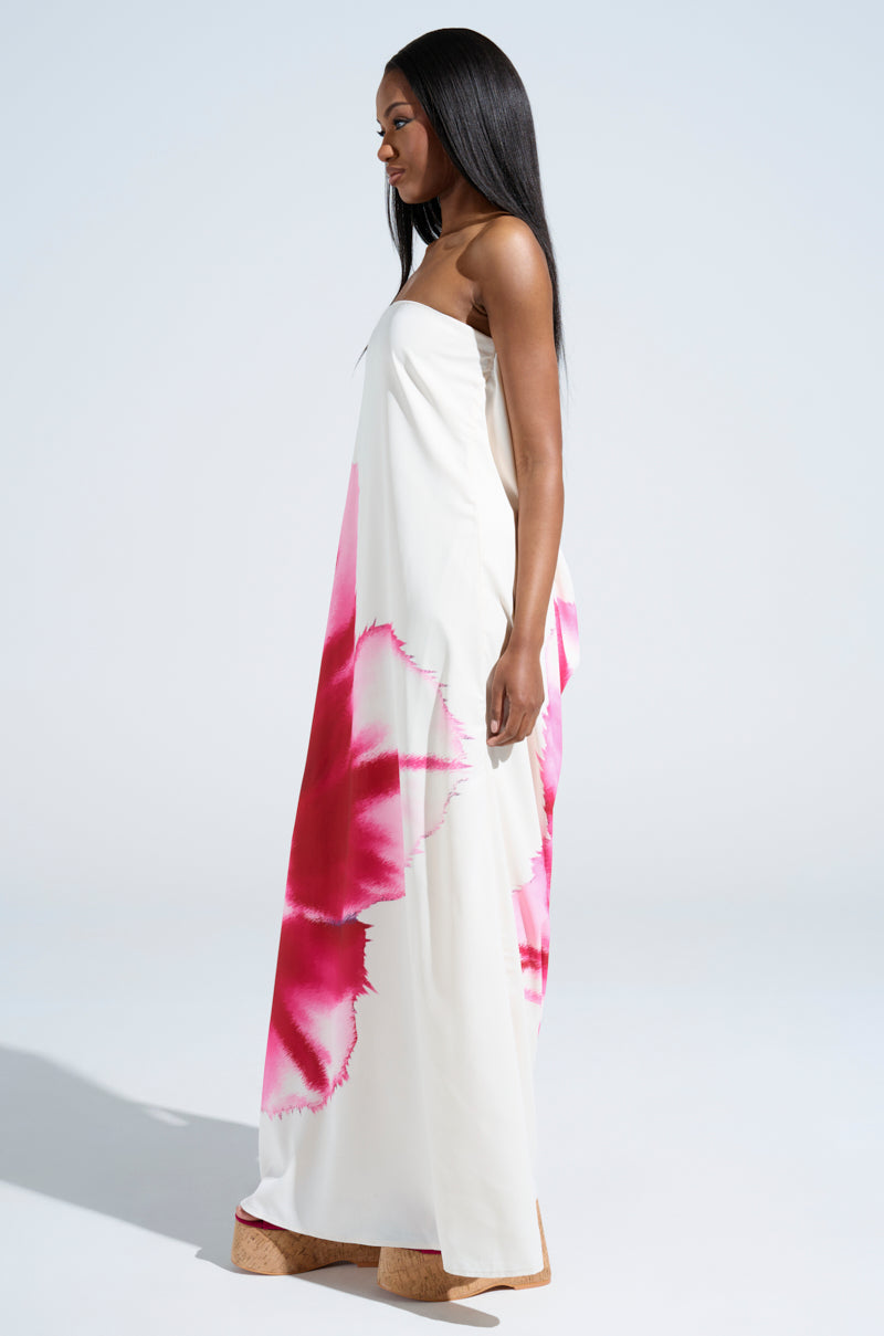 A woman with long straight hair and tan wedge sandals stands sideways in the SWEET AND SPICY SATIN MAXI DRESS, featuring bold pink tie-dye patterns and a plunging cowl back hemline, against a plain light background.