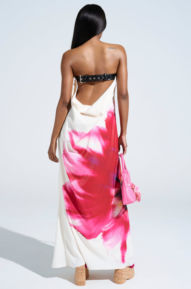 A woman with long straight hair, seen from behind, wears the SWEET AND SPICY SATIN MAXI DRESS with a plunging cowl back, bold pink and red floral print, tan platform shoes, and a small pink handbag.