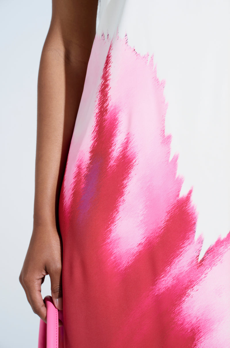 A person wears the SWEET AND SPICY SATIN MAXI DRESS—white satin with bold pink and red brushstroke pattern, featuring a plunging cowl back hem—holding a pink rectangular object, shown from shoulder to thigh.