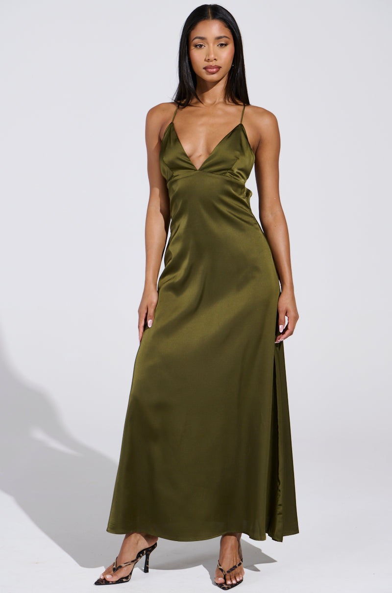 A woman stands elegantly in a STRAIGHT FROM THE 90'S satin maxi dress with thin straps, featuring a plunging neckline and flowing olive green fabric. She pairs it with black heeled sandals, her hair cascading freely against the plain white background.