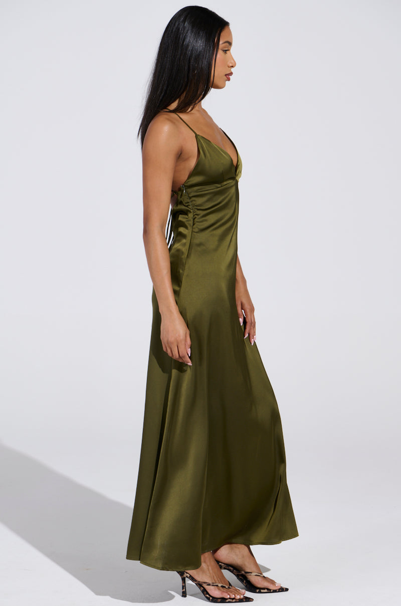 A person with long dark hair wears the STRAIGHT FROM THE 90'S satin maxi dress in olive green, featuring thin straps and a plunging neckline for a flowing silhouette. They pair it with high-heeled shoes and stand in profile against a neutral background.