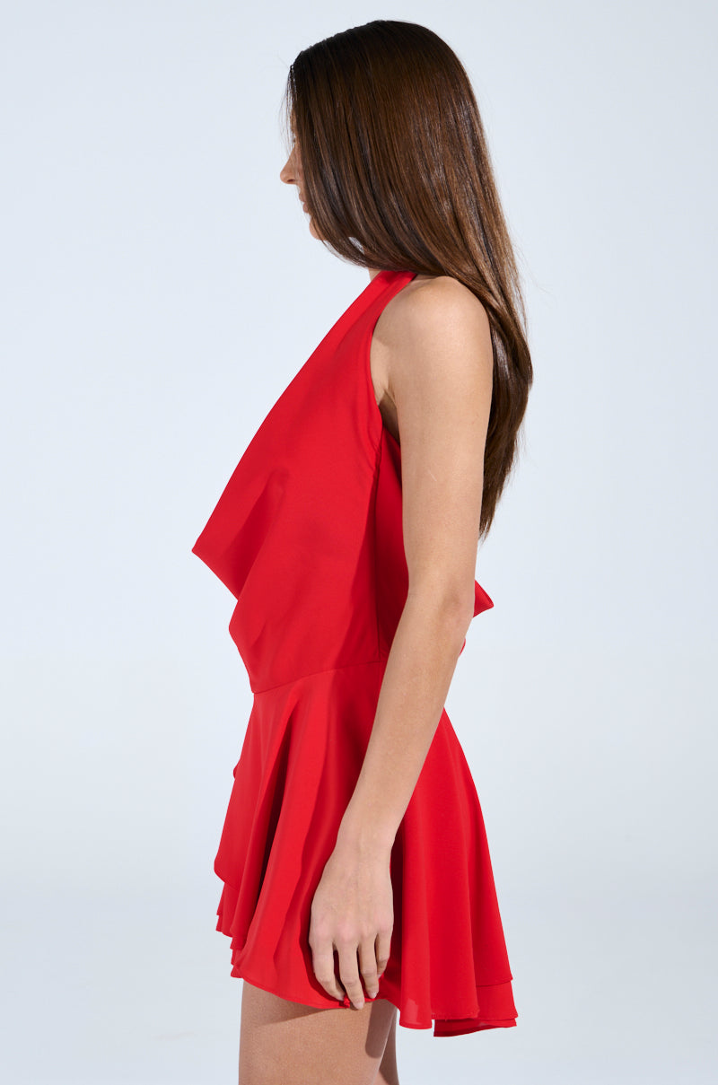 A woman with long brown hair stands sideways in a sleeveless ALL ABOUT ME MINI DRESS IN RED. The dress is flowing and features an open-back design against a plain white background.