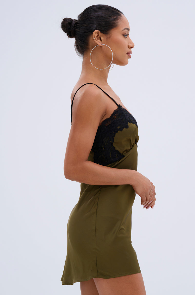 A woman in profile models the LET'S GET OUT OF HERE SATIN MINI DRESS, a sleeveless olive green mini with black lace trim, plunging V-neckline, and adjustable spaghetti straps. She pairs it with large hoop earrings and a sleek bun against a light background.