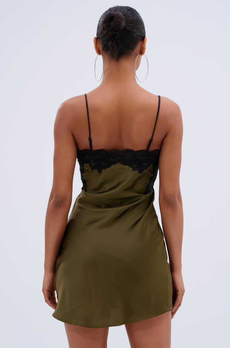 A woman with her hair in a bun and large hoop earrings models the LET'S GET OUT OF HERE SATIN MINI DRESS in olive green, featuring black lace trim and adjustable spaghetti straps, against a plain light background.
