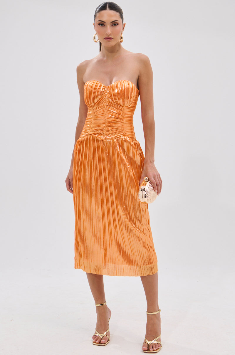A woman stands against a plain background wearing the ALL ABOUT ME SATIN BUBBLE MIDI DRESS IN ORANGE with a structured bodice and metallic sheen, styled with a small clutch, gold sandals, hoop earrings, and her hair tied back.
