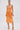 A woman stands against a plain background wearing the ALL ABOUT ME SATIN BUBBLE MIDI DRESS IN ORANGE with a structured bodice and metallic sheen, styled with a small clutch, gold sandals, hoop earrings, and her hair tied back.