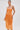 A woman wears the ALL ABOUT ME SATIN BUBBLE MIDI DRESS IN ORANGE, featuring shiny, strapless satin with a ruched, structured bodice. She holds a matching round clutch, stands against a plain background, and has her hair pulled back.