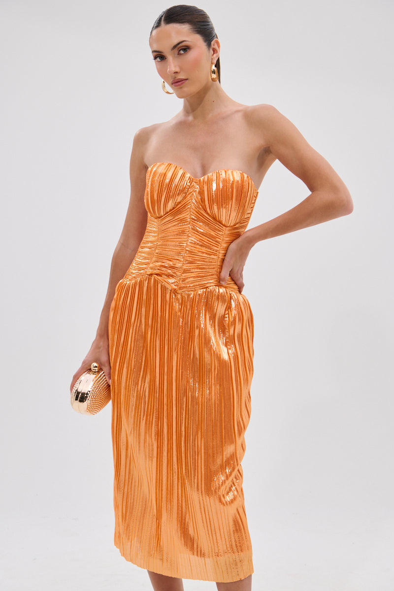 Woman poses in the AKIRA Label ALL ABOUT ME SATIN BUBBLE MIDI DRESS IN ORANGE, a strapless, metallic orange pleated dress featuring a structured bodice and shimmering sheen. She accessorizes with a gold clutch and hoop earrings against a light gray background.