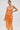 Woman poses in the AKIRA Label ALL ABOUT ME SATIN BUBBLE MIDI DRESS IN ORANGE, a strapless, metallic orange pleated dress featuring a structured bodice and shimmering sheen. She accessorizes with a gold clutch and hoop earrings against a light gray background.
