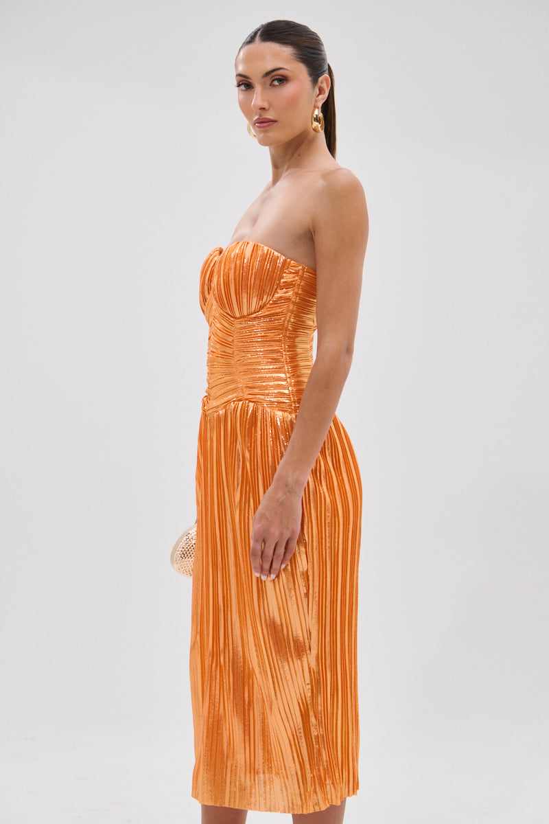 A woman stands sideways in the ALL ABOUT ME SATIN BUBBLE MIDI DRESS IN ORANGE, a shiny strapless pleated dress with a fitted bodice. She holds a small clutch, wears gold earrings, and has her dark hair pulled back against a light background.