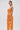 A woman stands sideways in the ALL ABOUT ME SATIN BUBBLE MIDI DRESS IN ORANGE, a shiny strapless pleated dress with a fitted bodice. She holds a small clutch, wears gold earrings, and has her dark hair pulled back against a light background.