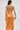 A woman faces away wearing the ALL ABOUT ME Satin Bubble Midi Dress in Orange, a strapless pleated style with a metallic sheen. She holds a matching clutch, sports a high ponytail, and completes the look with large round earrings.