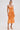 A woman models the ALL ABOUT ME SATIN BUBBLE MIDI DRESS IN ORANGE, styled with gold strappy heels, gold hoop earrings, and a textured clutch, standing against a plain white background.