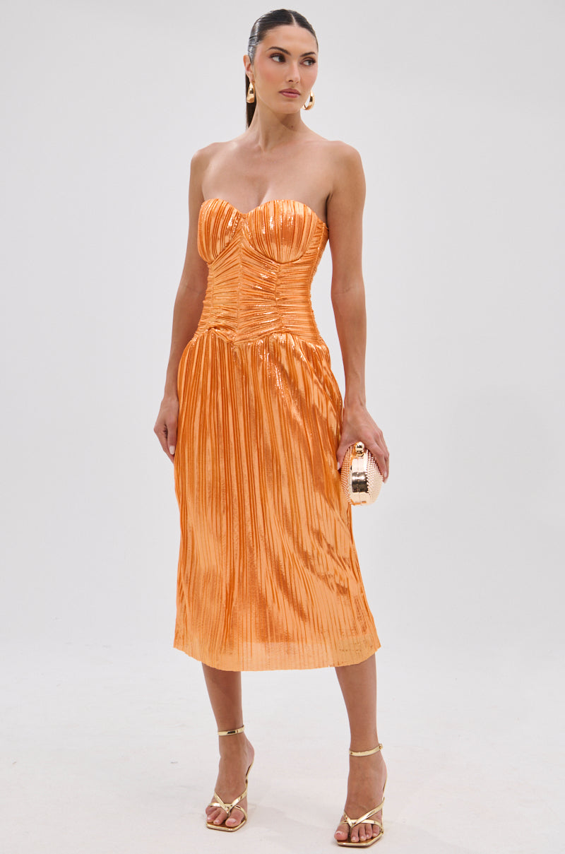 A woman in the ALL ABOUT ME SATIN BUBBLE MIDI DRESS IN ORANGE with a ruched bodice and metallic sheen stands against a plain backdrop, accessorizing with a small clutch, gold strappy heels, drop earrings, and sleek pulled-back hair.