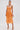 A woman in the ALL ABOUT ME SATIN BUBBLE MIDI DRESS IN ORANGE with a ruched bodice and metallic sheen stands against a plain backdrop, accessorizing with a small clutch, gold strappy heels, drop earrings, and sleek pulled-back hair.