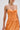 A woman wears the ALL ABOUT ME Satin Bubble Midi Dress in orange, featuring a strapless silhouette and metallic sheen. She holds a matching clutch, accessorizes with large gold hoops, and tucks her straight hair behind one ear.
