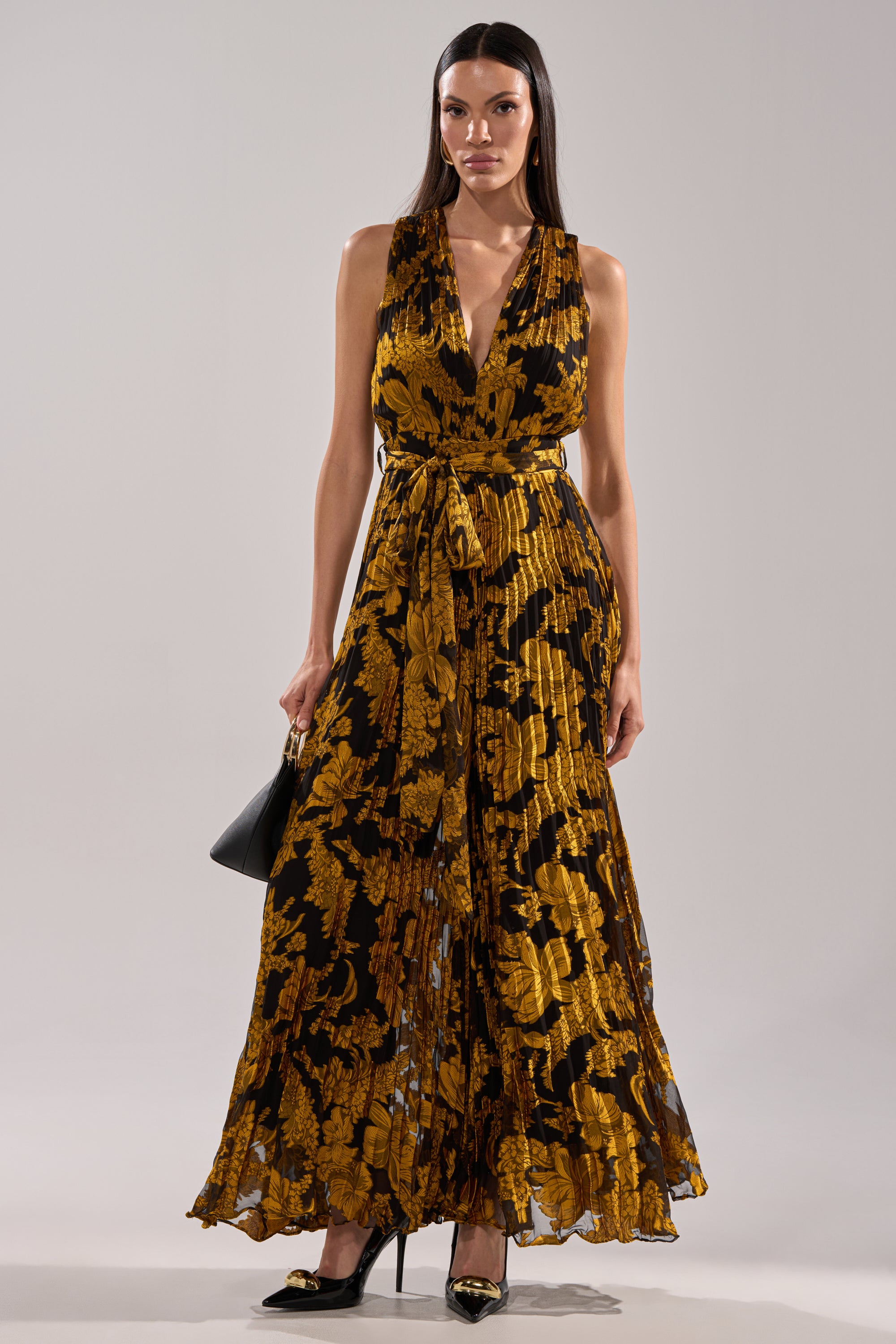 A woman wears the REGINA PRINTED WIDE LEG JUMPSUIT, a sleeveless, floor-length piece with gold floral patterns, a V-neckline, and waist belt. She accessorizes with a black handbag and black pointed-toe heels.