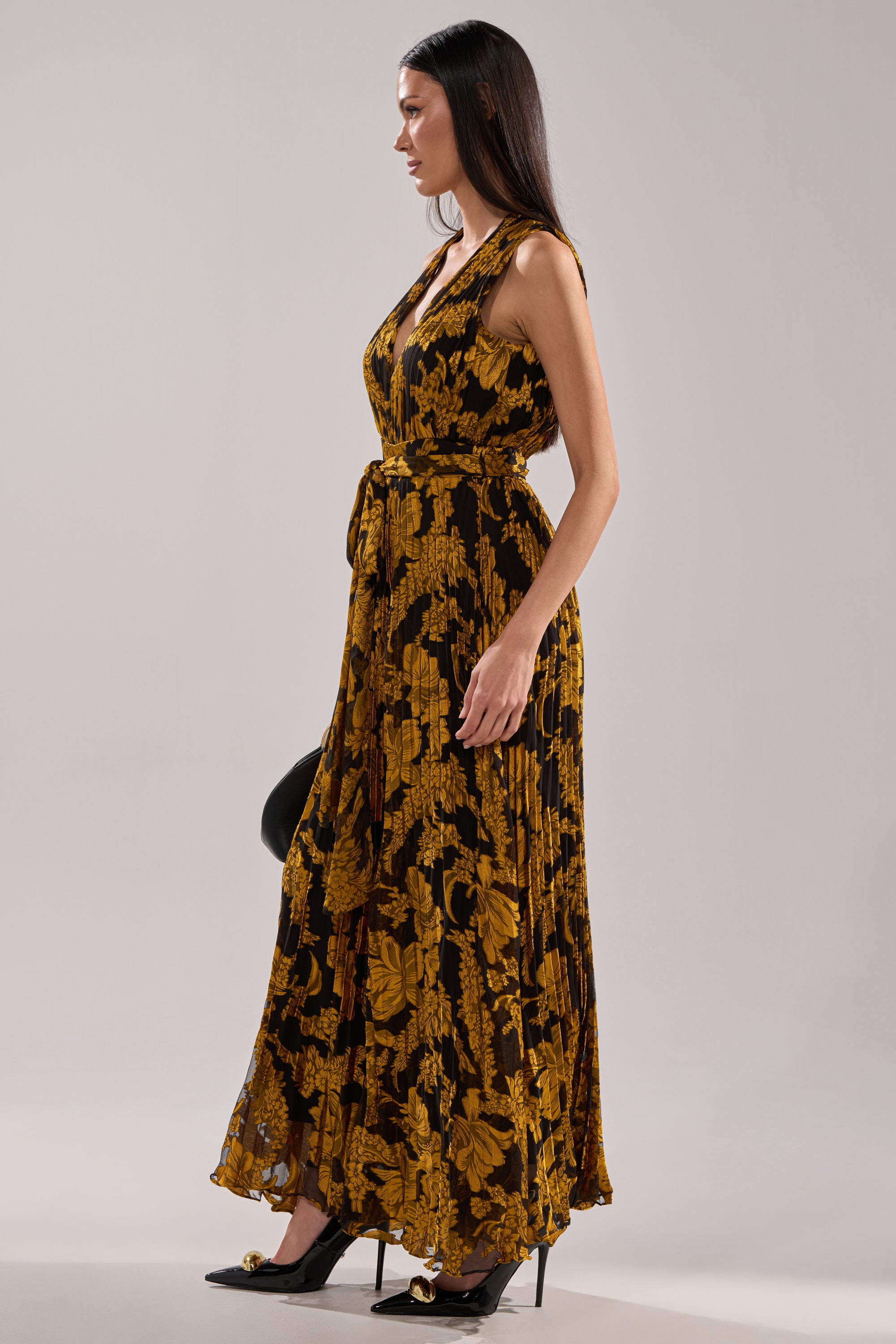 A woman in a sleeveless, floor-length AKIRA Label REGINA PRINTED WIDE LEG JUMPSUIT with bold yellow floral patterns, a matching belt, and black high heels stands in profile against a plain light background.