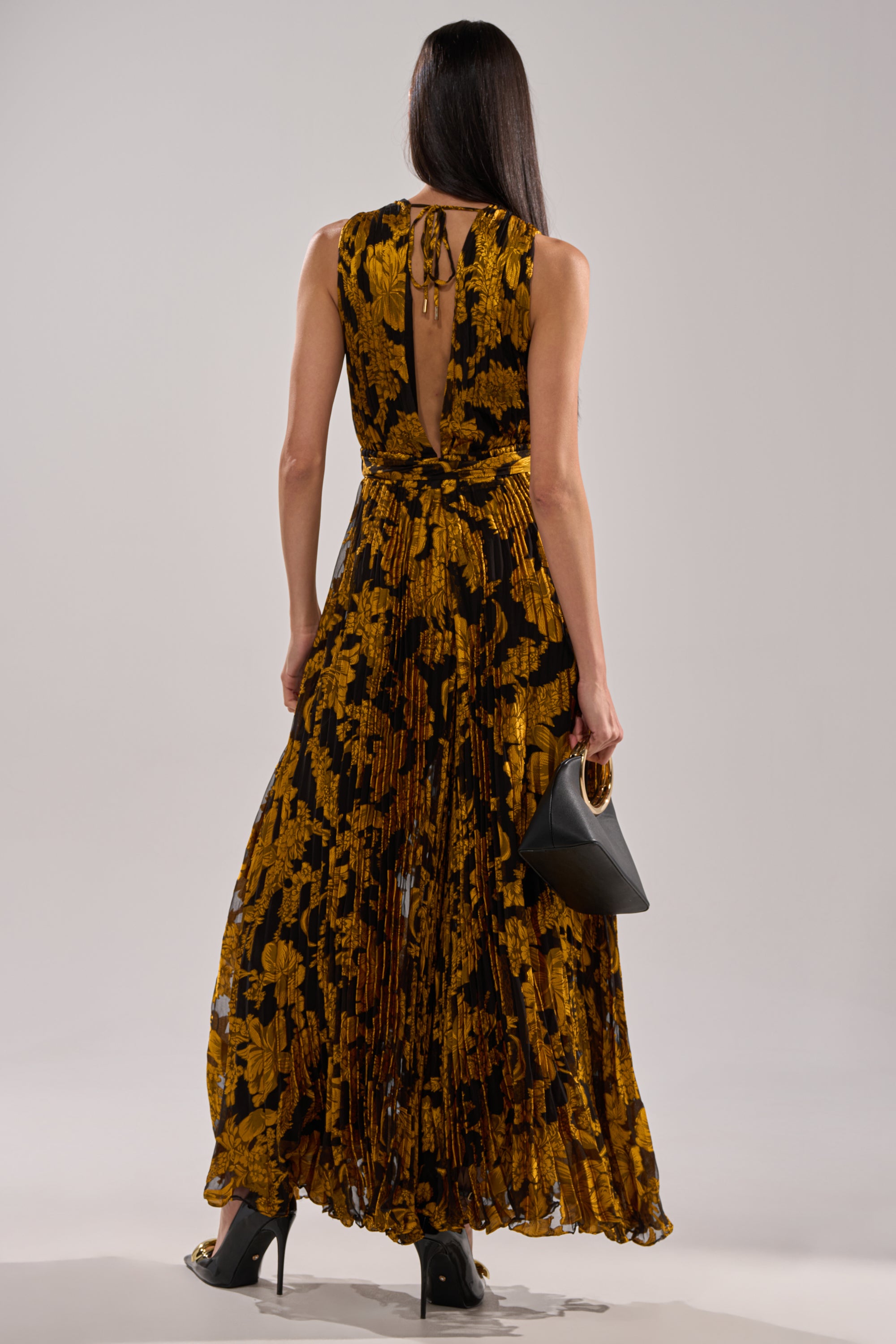 A woman with long dark hair, seen from behind, wears the REGINA PRINTED WIDE LEG JUMPSUIT in black and gold floral print with black heels and a small black handbag, standing against a plain background.
