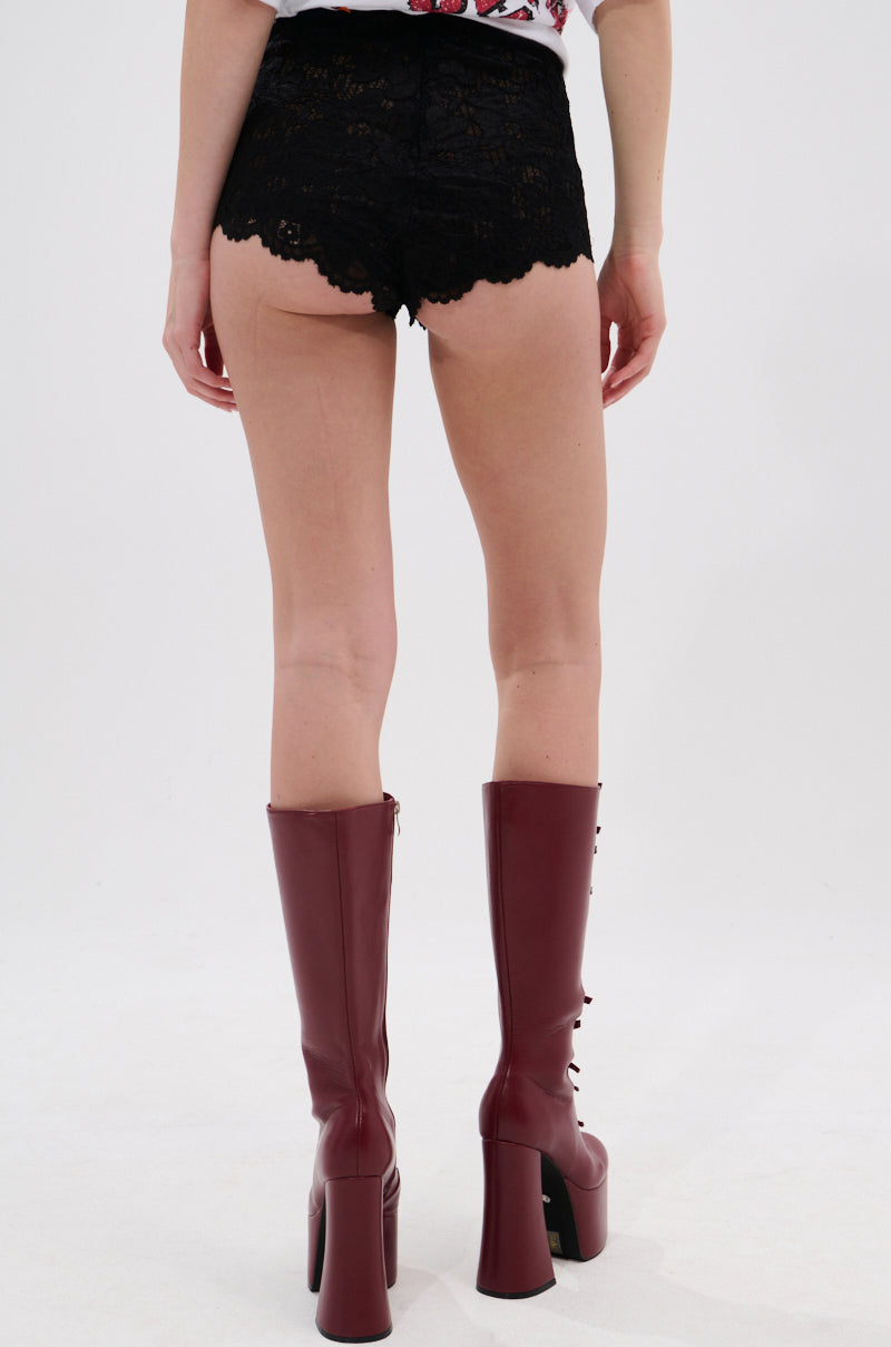 A person stands with their back to the camera, wearing TOO HOT TO HANDLE LACE HOT SHORT black shorts with a cheeky cut and tall, dark red platform boots against a plain white background. 4