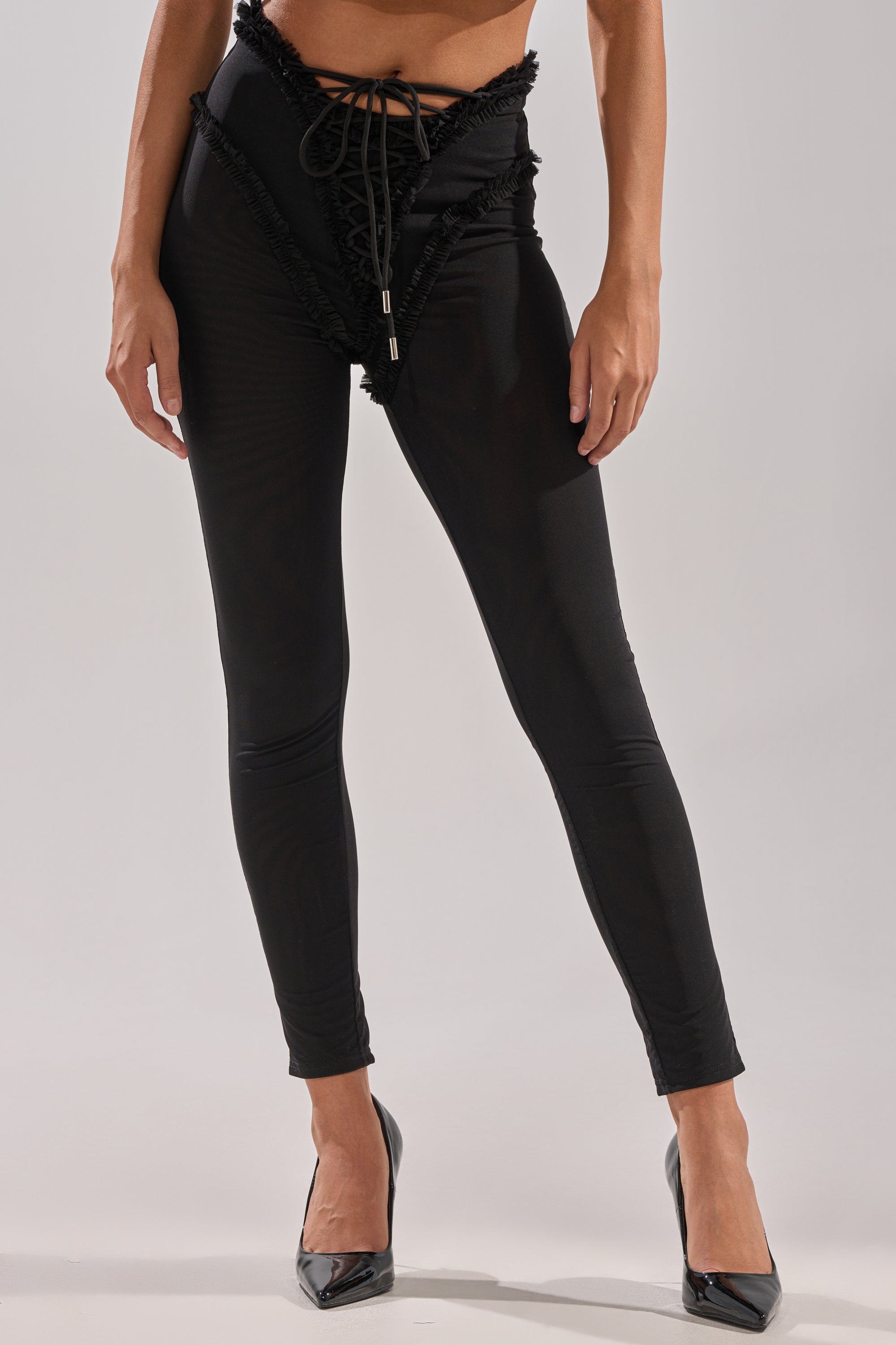 Wearing the KELLY MESH LACE UP LEGGING featuring high-rise mesh, lace-up front, and ruffle trim, paired with black pointed heels, a person stands against a plain background. 0
