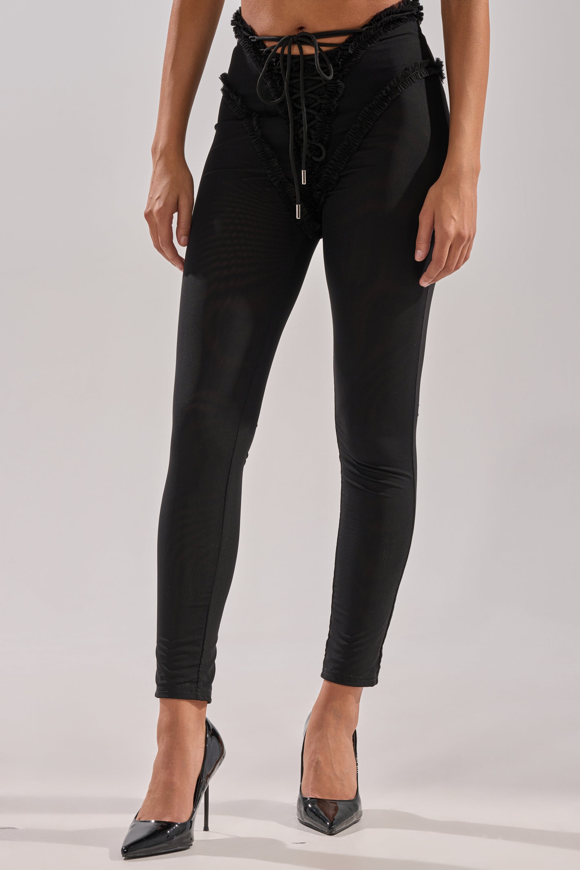 Wearing the KELLY MESH LACE UP LEGGING with ruffle details and black pointed high heels, a person stands against a plain light background. 1