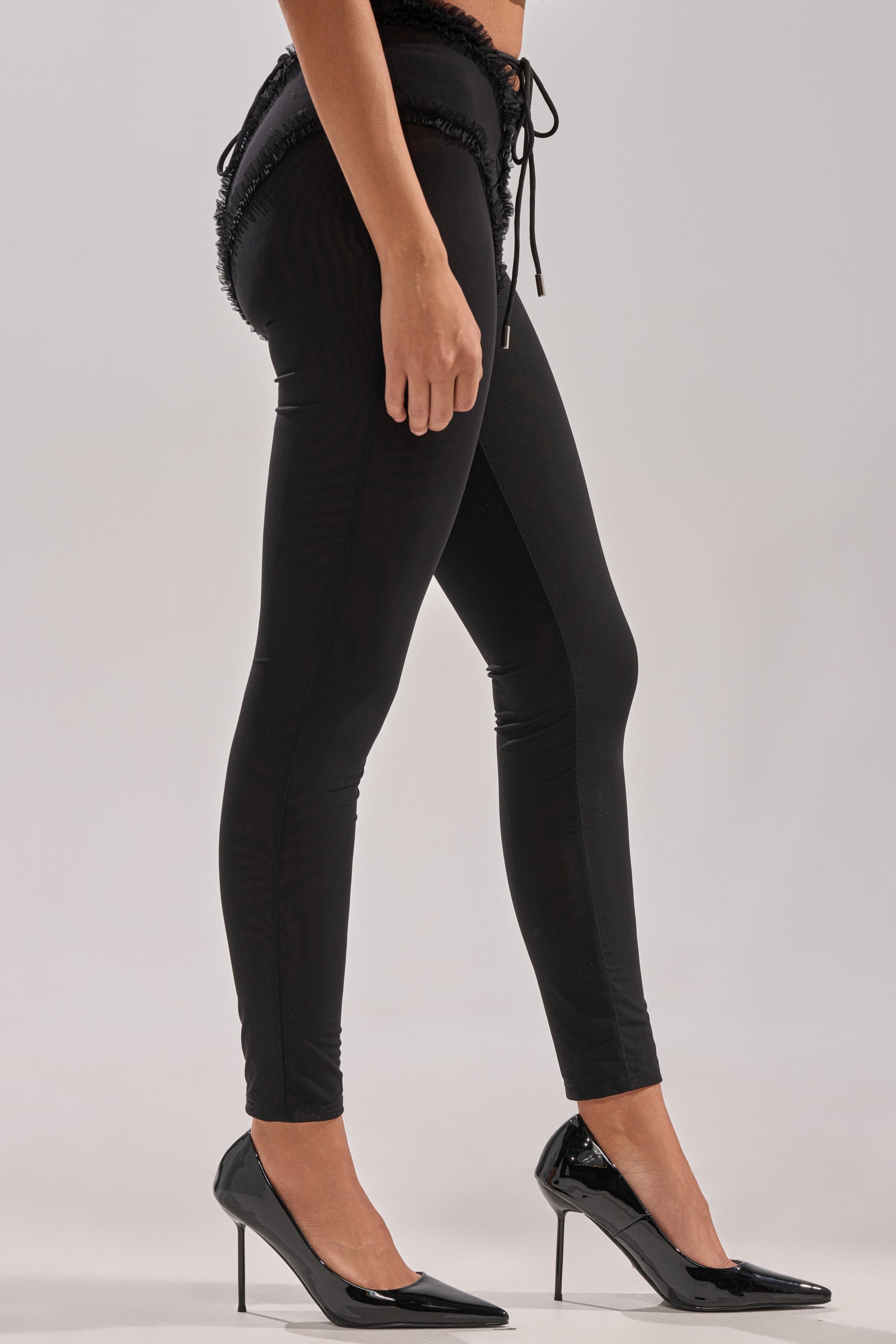A person stands sideways against a plain background, wearing the KELLY MESH LACE UP LEGGING and black pointed-toe high heels. 2