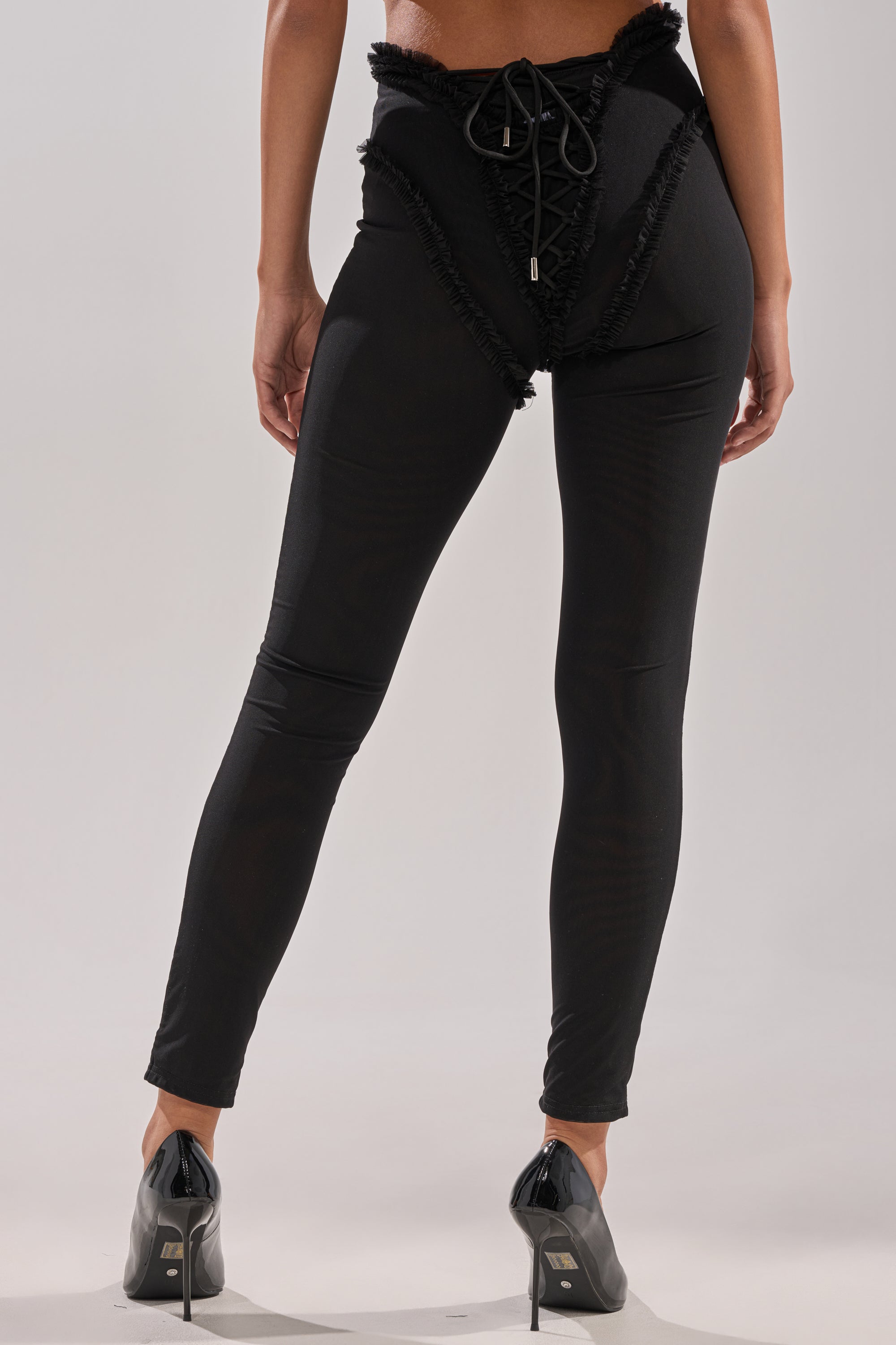 A person stands with their back to the camera, wearing the KELLY MESH LACE UP LEGGING featuring mesh lace-up details and ruffle trim at the waist, paired with black high-heeled shoes. 3