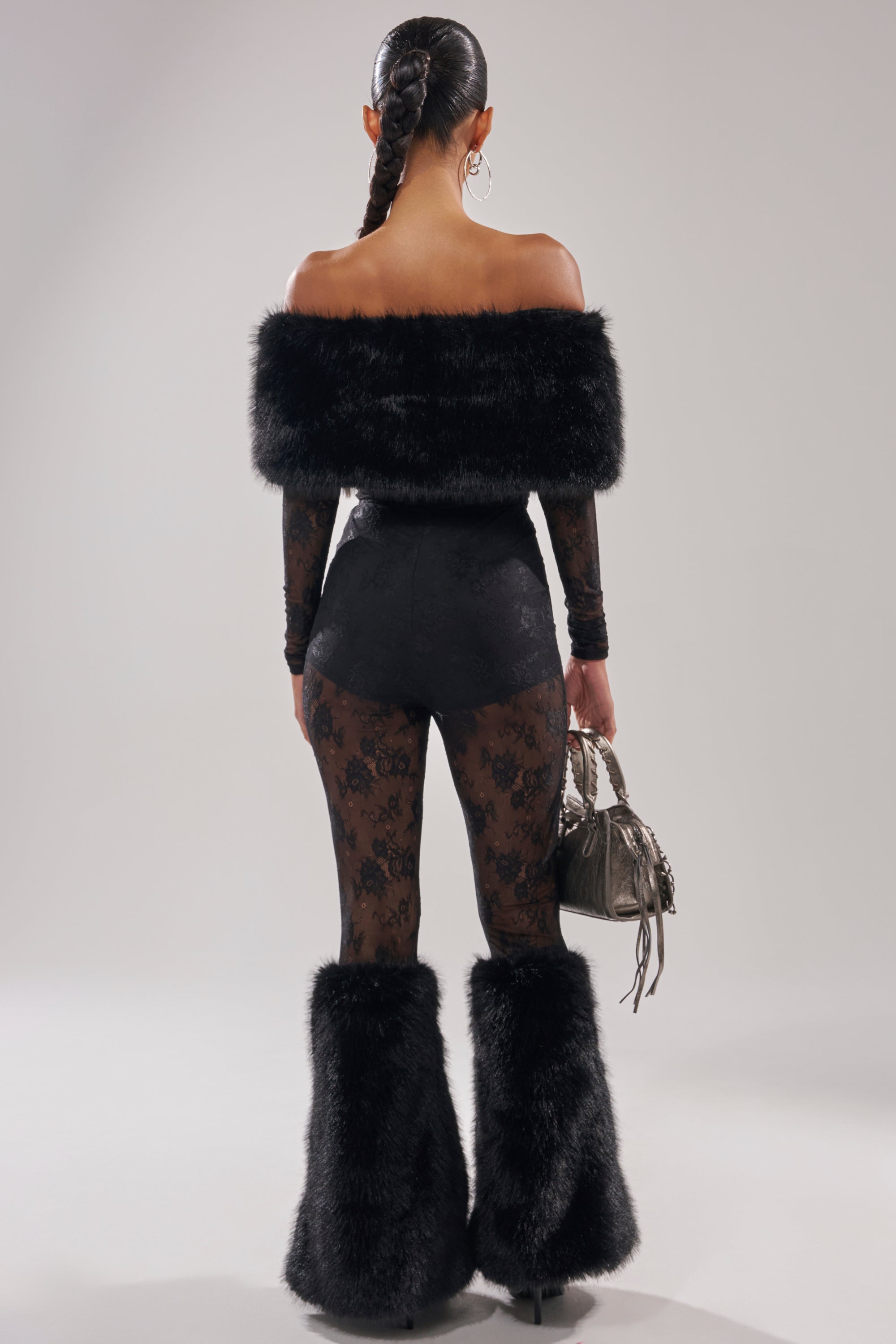 A woman faces away wearing the DELARA FUR TRIM LACE JUMPSUIT—an off-shoulder style with sheer black lace pants, fur trim, and faux fur boot covers. She carries a metallic handbag and has her hair in a tight braid with high heels.