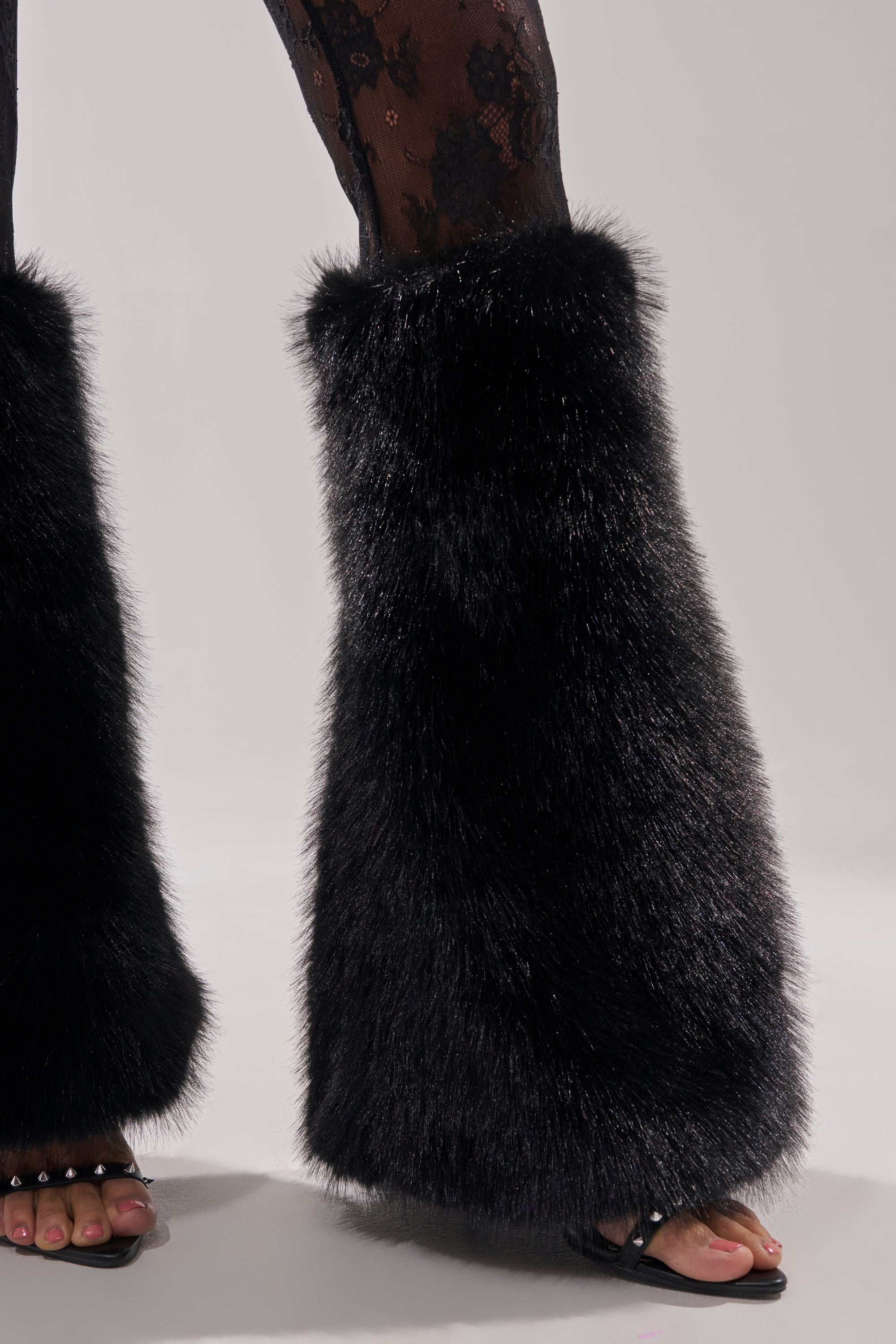 Wearing the DELARA FUR TRIM LACE JUMPSUIT, paired with black lace tights, fluffy black fur leg warmers over open-toe sandals, on a light surface.