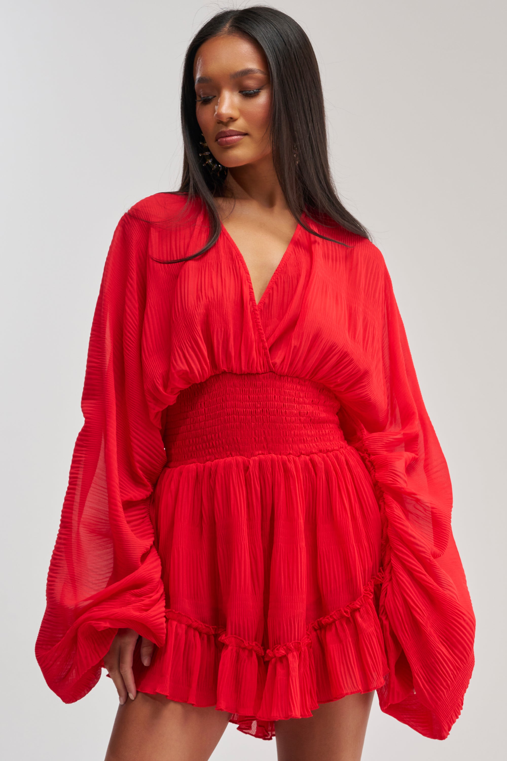 A woman poses against a plain light background, wearing the HOPELESS ROMANTIC ROMPER—a bright red mini romper with voluminous long sleeves and a ruffled hem, ideal for a fun date night look.